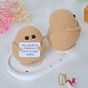 May include: Two crocheted potato figures in beige yarn, each with black button eyes. One potato holds a sign that reads "Life was all out of lemons, so here's a potato instead." The potatoes are on a white tray with a dotted design.