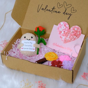 May include: A Valentine's Day gift box containing a crocheted bean figure holding a sign saying "You're My Favourite Human Bean." The box also includes a crocheted rose, a heart-shaped card, and various crocheted flowers. The box is filled with pink paper shreds.