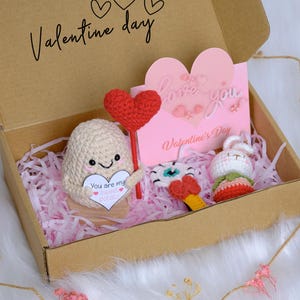 May include: A Valentine's Day gift box with crochet items. The box contains a crochet potato with a heart, a heart-shaped card, a crochet bunny, and other small crochet items. The box is filled with pink paper and has the words "Valentine day" on the lid.
