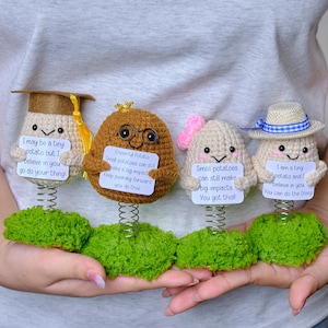 Puede incluir: Cuatro figuras de patata hechas a mano con ganchillo, con sombreros y letreros. Las patatas están unidas a muelles y bases verdes. Cada patata sostiene un letrero con un mensaje inspirador.