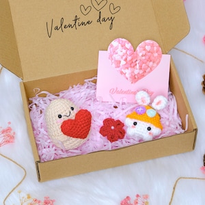 Puede incluir: Una caja de regalo de San Valentín con un corazón de ganchillo, un conejito de ganchillo y una flor roja. La caja contiene relleno de papel rosa y una tarjeta rosa en forma de corazón. La tapa de la caja marrón tiene las palabras "Valentine day".