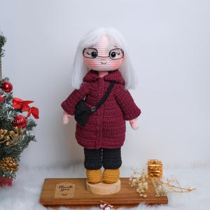 Könnte beinhalten: Handgefertigte Häkelpuppe einer Frau in einem weinroten Mantel, schwarzer Hose und gelben Stiefeln. Die Puppe hat weißes Haar, eine Brille und eine schwarze Handtasche. Sie steht auf einem Holzsockel, im Hintergrund eine kleine Geschenkbox und ein Weihnachtsbaum.