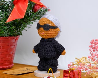 May include: A handmade crochet doll wearing sunglasses, a black suit, and a silver earring. The doll has gray hair and is standing on a wooden base. A small Christmas tree with a red bow and a gift box are in the background.