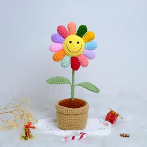 May include: A handmade crochet sunflower with a smiling yellow face and colorful petals. The flower sits in a brown pot with a green stem and leaves. The pot is made of a woven material. A cheerful, decorative item.