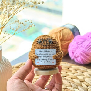 Könnte beinhalten: Eine braune, gehäkelte Kartoffelfigur mit einer schwarzen Drahtbrille, die auf einem kleinen Holzsockel steht. Sie hält ein weißes, rechteckiges Schild mit der Aufschrift: Cheerful Potato, Small potatoes can still make a big impact! Keep pushing forward!
