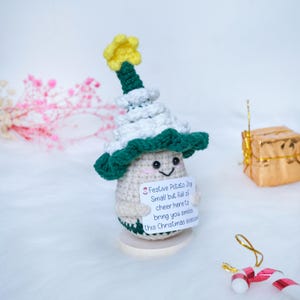 Op de afbeelding: Een gehaakte aardappel figuur met een wit en groene kerstboom hoed met een gele ster. De aardappel houdt een bord vast met de tekst "Festive Potato Joy Small but full of cheer here to bring you smiles this Christmas season."