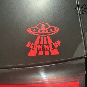 Pode incluir: Adesivo de vinil vermelho com um OVNI com uma figura alienígena dentro, emitindo luz. O texto "BEAM ME UP" está abaixo do OVNI. O adesivo é aplicado em uma superfície preta.