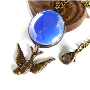 May include: A blue glass pendant with a bronze bird charm hangs from a bronze chain with a lobster clasp.