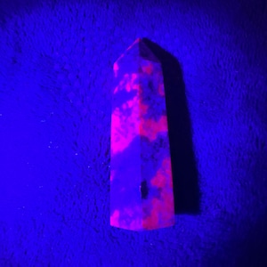 Puede incluir: Una punta de cristal rosa y púrpura con un patrón nublado y veteado. El cristal está iluminado con una luz negra, lo que hace que brille intensamente.