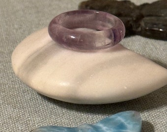 May include: A purple amethyst ring with a smooth, polished finish. The ring is displayed on a white stone surface.