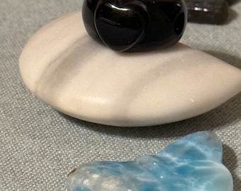 Può includere: Un anello nero a forma di cuore in ossidiana lucidata. L'anello è appoggiato su una roccia bianca con una pietra blu a forma di farfalla sullo sfondo.