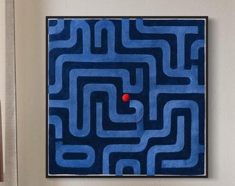 May include: A square wall art piece featuring a dark blue maze pattern on a navy background with a single small red sphere positioned near the center. The artwork is framed in a thin black border and hangs on a plain wall above a white sofa.