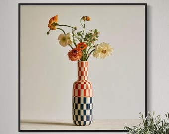 May include: A square framed wall art print featuring a tall cylindrical vase with a checkered pattern in red and white on the top half and blue and white on the bottom half, containing a bouquet of orange, white, and yellow ranunculus flowers.
