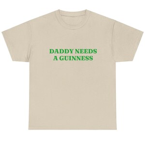 May include: A beige t-shirt with the text "DADDY NEEDS A GUINNESS" in green block letters. The shirt has a crew neck and short sleeves. The text is centered on the front of the shirt.