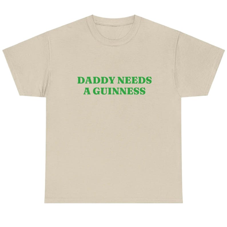 May include: A beige t-shirt with the text "DADDY NEEDS A GUINNESS" in green block letters. The shirt has a crew neck and short sleeves. The text is centered on the front of the shirt.