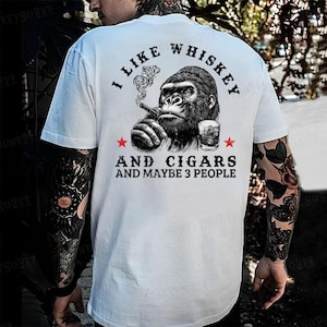 May include: White t-shirt with a graphic of a gorilla smoking a cigar and drinking whiskey. The text reads "I LIKE WHISKEY AND CIGARS AND MAYBE 3 PEOPLE" in black font. Two red stars flank the text.