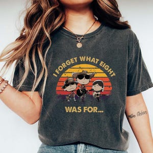 May include: Dark gray t-shirt featuring a cartoon band playing guitars against a sunset backdrop. The text "I FORGET WHAT EIGHT WAS FOR..." is printed above the band. The shirt has a vintage, worn look.