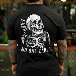 Puede incluir: Camiseta negra con un dise&ntilde;o de esqueleto blanco. El esqueleto tiene un dedo en la boca, con las palabras "SHHH" arriba y "NO ONE CARES" abajo. La camiseta es de manga corta.