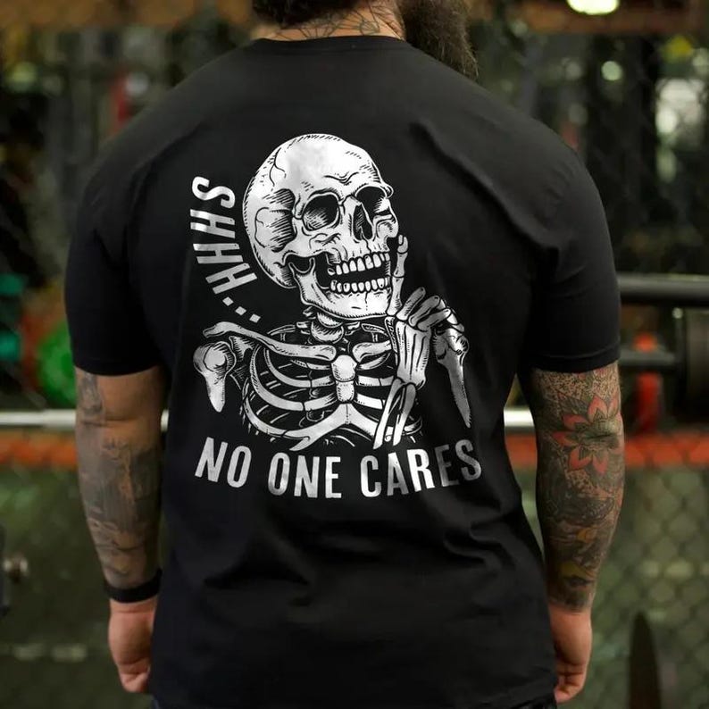 Puede incluir: Camiseta negra con un dise&ntilde;o de esqueleto blanco. El esqueleto tiene un dedo en la boca, con las palabras "SHHH" arriba y "NO ONE CARES" abajo. La camiseta es de manga corta.