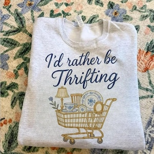 May include: Light gray sweatshirt with the text "I'd rather be Thrifting" in navy blue script. A gold shopping cart graphic is filled with vintage items like a lamp, dishes, and jewelry.