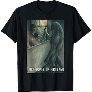 May include: Black t-shirt featuring a fantasy illustration. The artwork depicts a person touching a werewolf through a window. The text "THEY DON'T UNDERSTAND" is printed below the image. The shirt is made of a soft material.