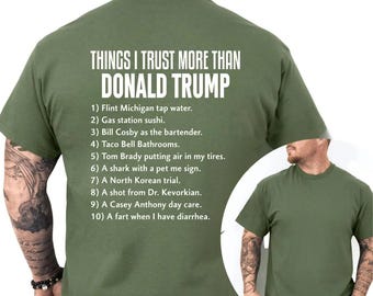 May include: Olive green t-shirt with white text. The text reads "THINGS I TRUST MORE THAN DONALD TRUMP" followed by a list of humorous comparisons. The shirt is a crew neck style.