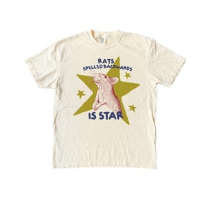 May include: Cream-colored t-shirt featuring a rat illustration inside a green star. The text reads "RATS SPELLED BACKWARDS IS STAR" in blue. The design includes small yellow stars.