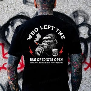 May include: Black t-shirt with a graphic of a gorilla smoking a cigar and a glass of whiskey. The text reads "WHO LEFT THE BAG OF IDIOTS OPEN SERIOUSLY! THEY'RE EVERYWHERE!" in white lettering.