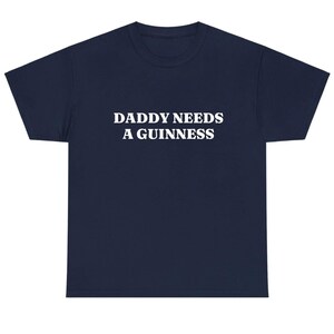 May include: Navy blue t-shirt with the white text "DADDY NEEDS A GUINNESS" printed across the front. The shirt has a crew neck and short sleeves. A casual, comfortable tee.