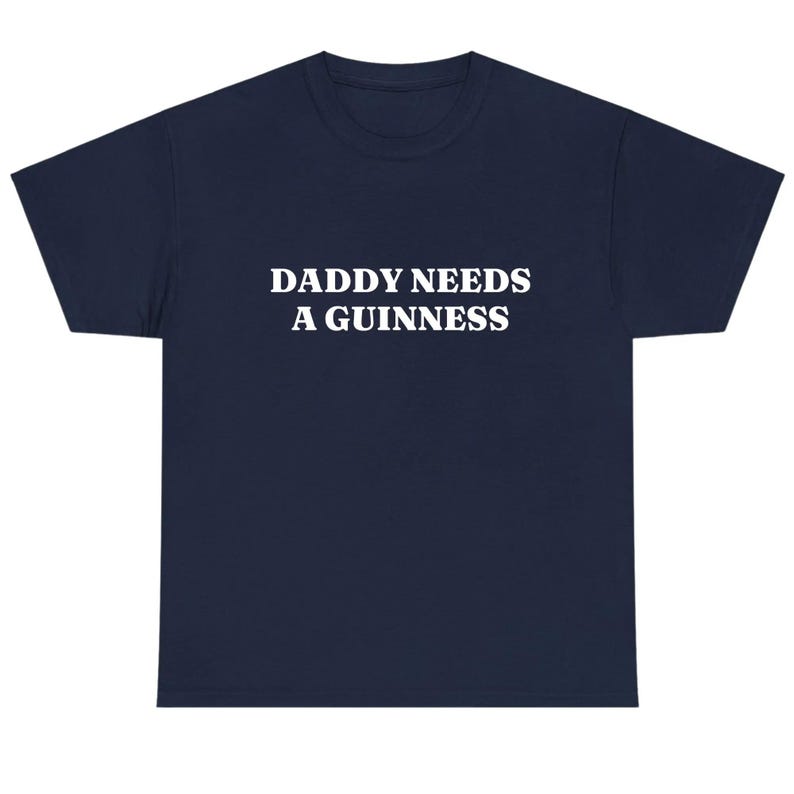 May include: Navy blue t-shirt with the white text "DADDY NEEDS A GUINNESS" printed across the front. The shirt has a crew neck and short sleeves. A casual, comfortable tee.