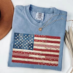 May include: A light blue t-shirt with a patchwork American flag design. The flag features a blue field with white stars and red and white striped sections. The shirt has a Comfort Colors tag and a gold necklace.