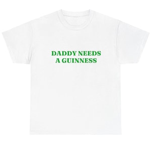 May include: White t-shirt with the green text "DADDY NEEDS A GUINNESS" across the chest. The shirt is a casual, crew neck style, suitable for everyday wear. The text is in a bold, sans-serif font.