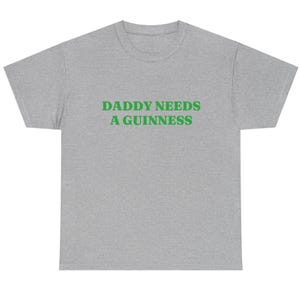 May include: A heather gray t-shirt with the text "DADDY NEEDS A GUINNESS" in green block letters. The shirt is a casual style, suitable for everyday wear.