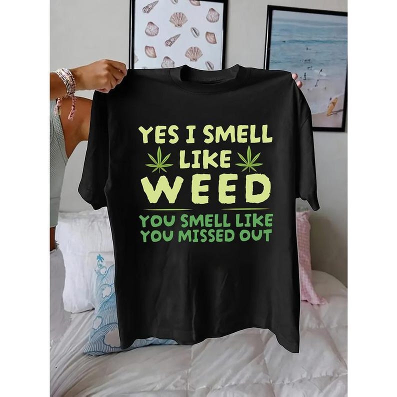 Op de afbeelding: Zwart T-shirt met de tekst "YES I SMELL LIKE WEED" in geel en groen. Daaronder twee cannabisbladeren en de zin "YOU SMELL LIKE YOU MISSED OUT" in groen.