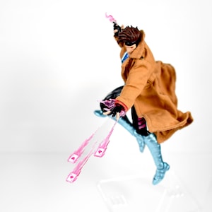 May include: Action figure of a character in a brown coat, black and blue outfit, and a mask. The figure is mid-jump, with pink energy effects emanating from the hands and a playing card design. The figure is on a clear stand.