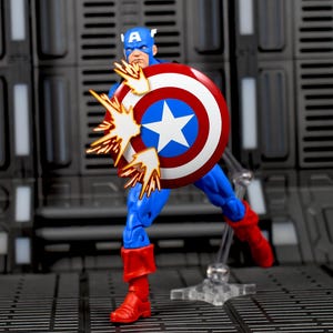 May include: A Captain America action figure in a dynamic pose, holding his iconic shield. The shield is red, white, and blue with a white star. The figure is wearing a blue suit, red boots, and a blue helmet. Explosive effects are emanating from the shield.