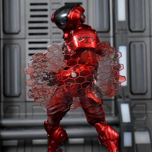 May include: A red action figure in a futuristic suit with a black helmet and a clear, honeycomb-patterned shield. The figure is posed against a gray, industrial-style background, showcasing its metallic finish and intricate design.