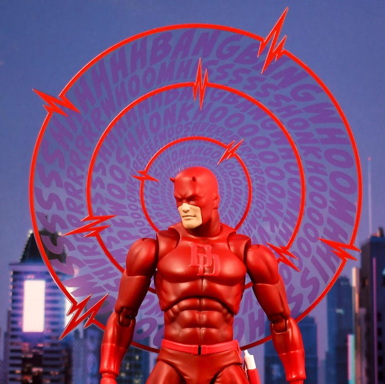 K&ouml;nnte beinhalten: Eine rote und schwarze Daredevil-Actionfigur mit einer roten Maske und einem roten Anzug. Die Figur steht vor einem wirbelnden Hintergrund mit den Worten "Bang", "Whoom", "Ssh" und "Honk", die in einem Kreis wiederholt werden.