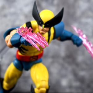 May include: A Wolverine action figure in a dynamic pose, featuring a yellow and blue costume with a red belt. The figure has black headgear and is wielding pink energy claws, creating a sense of action and power.