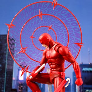 May include: A red action figure of Daredevil, a superhero known for his heightened senses, is posed in a dynamic stance. He is holding a white stick in his right hand and a red billy club in his left hand. The figure is set against a background of a city skyline with a purple and red comic book sound effect graphic behind him.