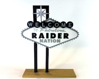Könnte beinhalten: Ein dekoratives Schild mit einer weißen rautenförmigen Tafel und dem schwarzen Text WELCOME TO Fabulous RAIDER NATION. Das Schild ist auf zwei schwarzen vertikalen Pfosten montiert, mit einem silbernen Stern an der Spitze und einem beigen Sockel.