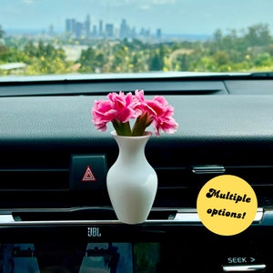 May include: A white ceramic vase with pink carnations inside, sitting on the dashboard of a car. The vase is a simple, elegant design and the flowers add a touch of color to the car's interior.  Multiple options!