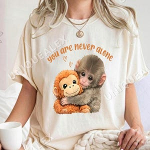 May include: Cream-coloured t-shirt with a graphic of two hugging monkeys. The orange monkey is hugging a grey monkey. The text "you are never alone" is printed above the monkeys in orange.