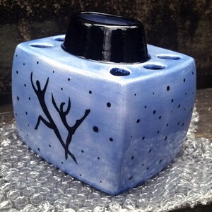 May include: A blue ceramic toothbrush holder with a black top and a design of two figures dancing. The holder has a speckled blue glaze and a white background.