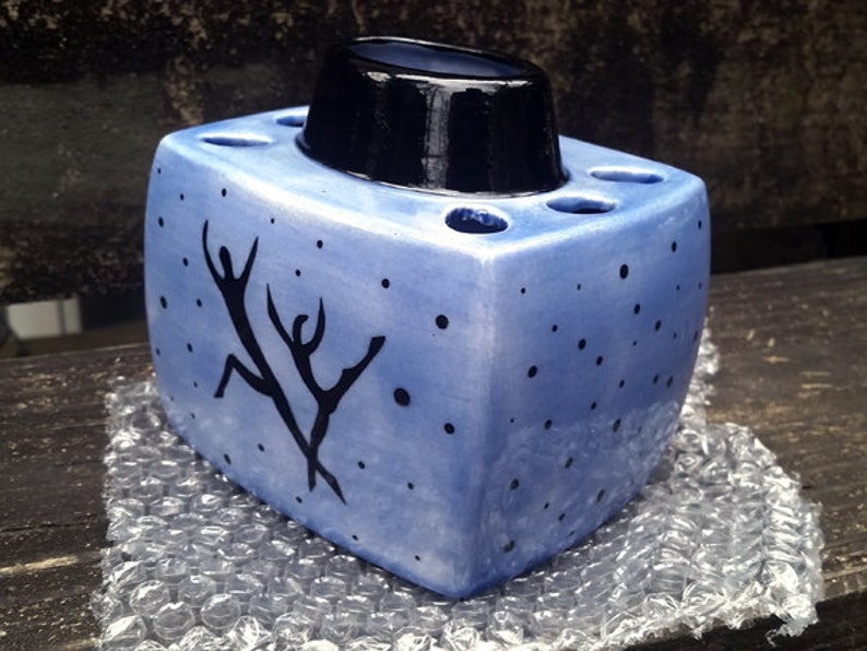 May include: A blue ceramic toothbrush holder with a black top and a design of two figures dancing. The holder has a speckled blue glaze and a white background.