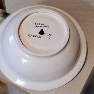 Op de afbeelding: Witte keramische kom met een zwarte inscriptie die "Terra Treasures" en "Cafes" luidt met een driehoeksymbool en het copyright-symbool en het jaar 2000.