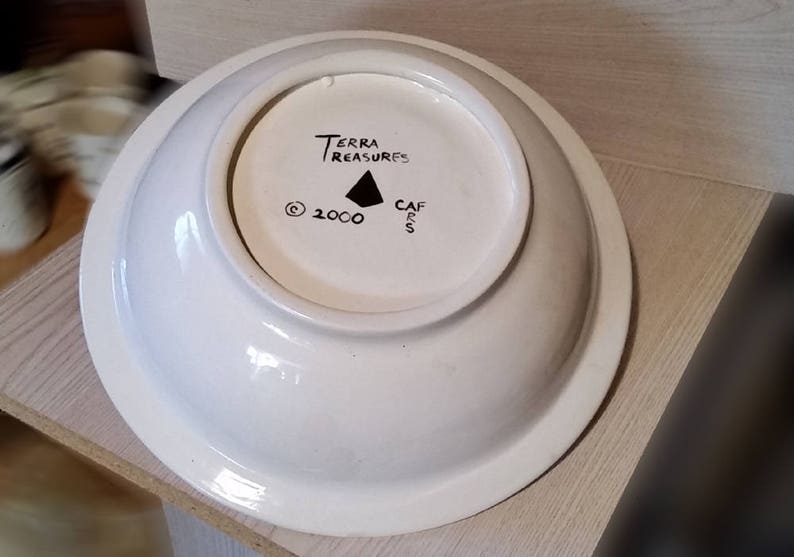 Op de afbeelding: Witte keramische kom met een zwarte inscriptie die "Terra Treasures" en "Cafes" luidt met een driehoeksymbool en het copyright-symbool en het jaar 2000.