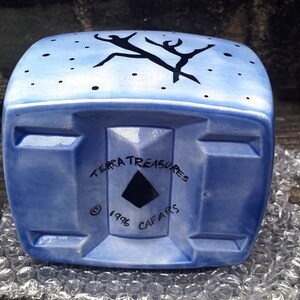 May include: A blue ceramic planter with a stylized black design of two figures dancing. The planter has a square shape with rounded edges and a flat bottom. The bottom of the planter has the text "Terra Treasures &copy; 1996 Cafairs".