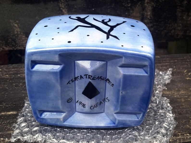 May include: A blue ceramic planter with a stylized black design of two figures dancing. The planter has a square shape with rounded edges and a flat bottom. The bottom of the planter has the text "Terra Treasures &copy; 1996 Cafairs".