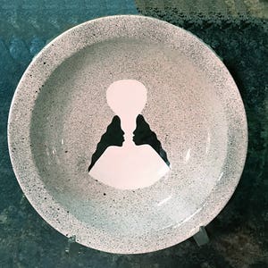 Op de afbeelding: Een witte keramische kom met een gespikkeld grijs glazuur. De kom heeft een zwart silhouet van twee mensen die elkaar aankijken, waardoor &eacute;&eacute;n figuur ontstaat.